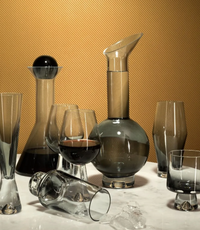 The Tom Dixon Tank Black Decanter is showcased with modern mouth-blown glassware, including tumblers and wine glasses, arranged on marble with a gold backdrop. One tumbler lies tipped over among scattered ice cubes.