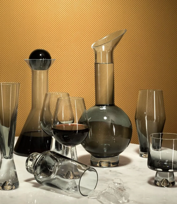 The Tom Dixon Tank Black Decanter is showcased with modern mouth-blown glassware, including tumblers and wine glasses, arranged on marble with a gold backdrop. One tumbler lies tipped over among scattered ice cubes.