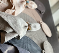 Whigstock Studio's Hand Dyed Linen Napkin Set of 4 in Pebble is folded on textured round placemats atop a dark table, each napkin held by a decorative ring. Soft light bathes the neutral-toned scene from the right.
