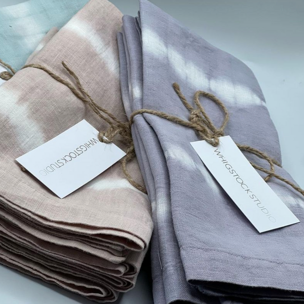 Two bundles of Whigstock Studio Hand Dyed Linen Napkin Set of 4 - Lavender Gray are tied with twine and feature subtle tie-dye patterns. Each sustainable set includes a white tag labeled Whigstock Studio.
