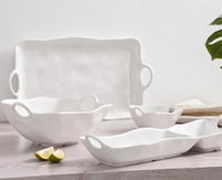The PAMPA BAY Ivy Melamine 2 Section Server, a white BPA-free piece with wavy edges and handles, is arranged on a light wooden surface beside bowls, trays, lime wedges, and a green plant partially visible to the right.