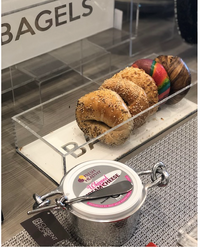 A row of assorted bagels is displayed in an Eye Candy Bagel Tray, labeled BAGELS, with whipped cream cheese and a serving knife in front on the counter.