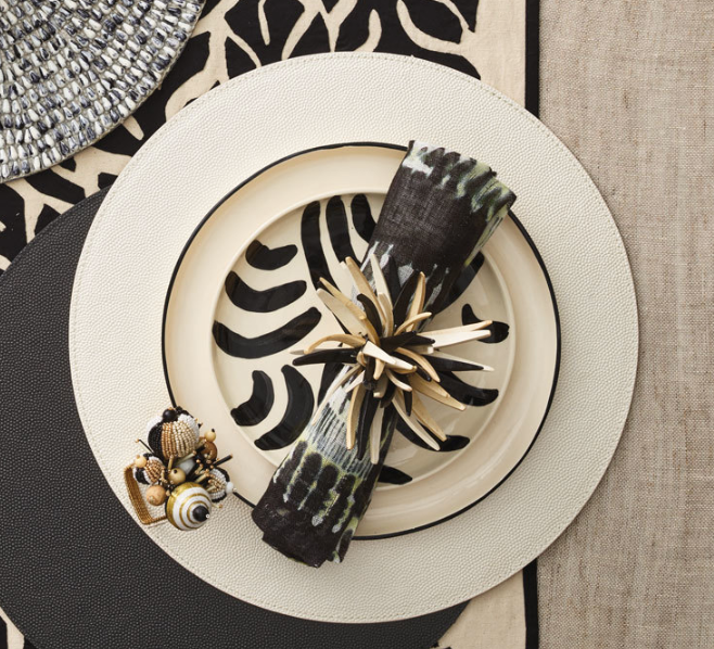 A decorative table setting with a cream plate featuring black and white leaves, a patterned black napkin in a gold ring, seashells, and textured Kim Seybert Shagreen Placemat White Set of 4 for an elegant dinner party.