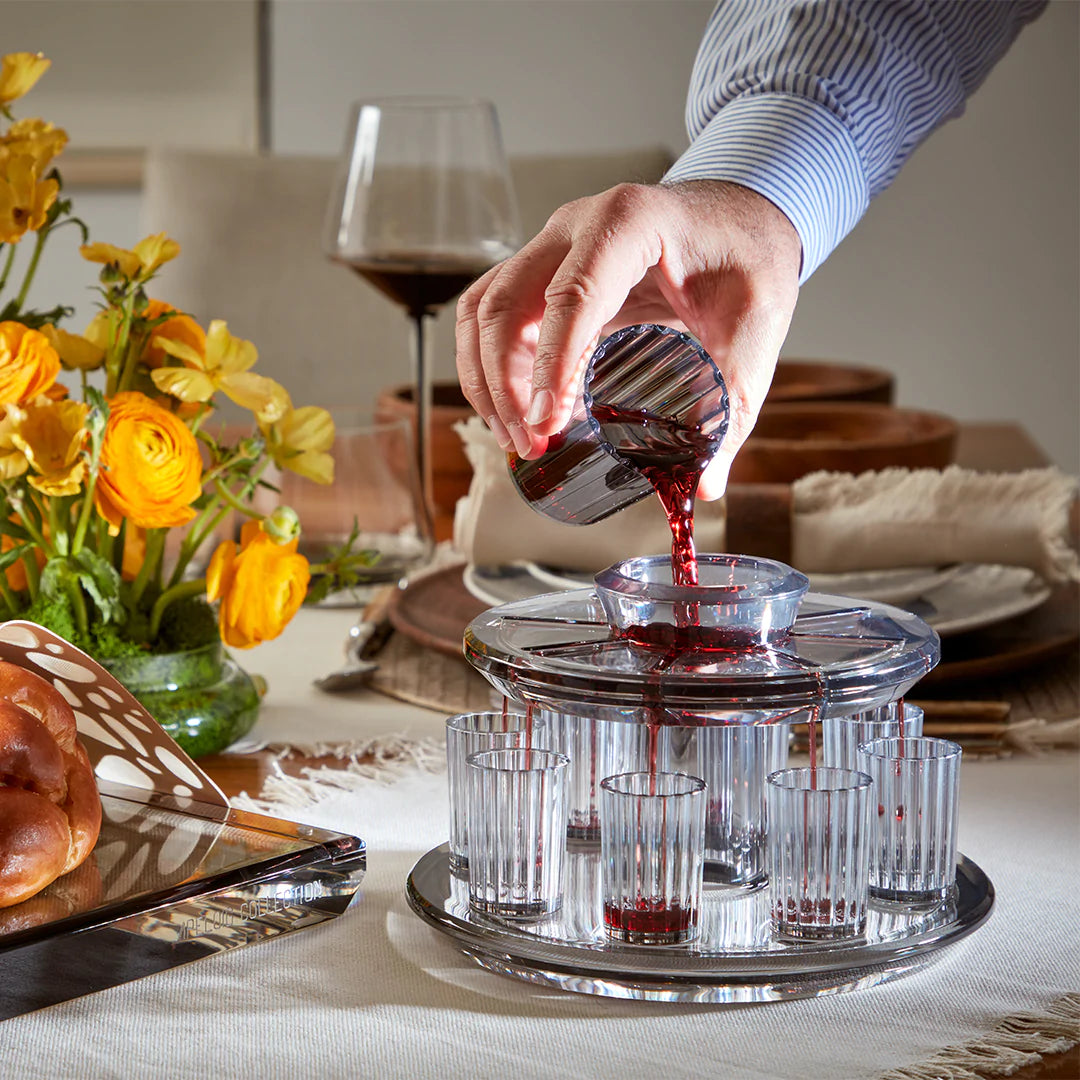 A person pours red wine into the Apeloig Kiddush Wine Fountain Set, which dispenses wine into several small glasses on a tray for a festive Judaica celebration, with flowers, bread, and wine glasses set on the table in the background.