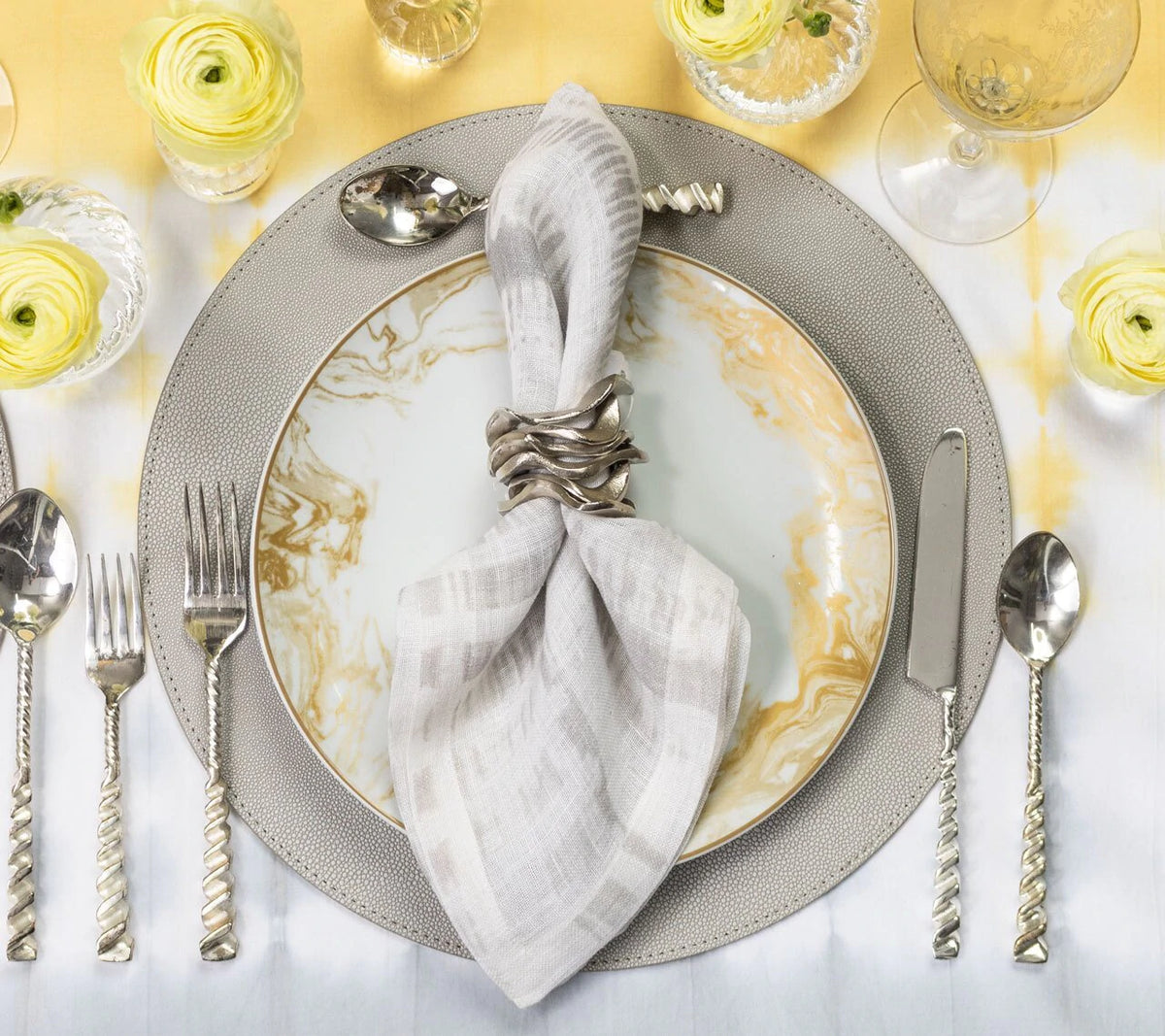 Elegant table setting featuring Kim Seybert's Shagreen Placemat in Elephant Grey, marbled plate, textured silver utensils, yellow flowers, and a light napkin with a silver ring over a yellow and white gradient tablecloth.