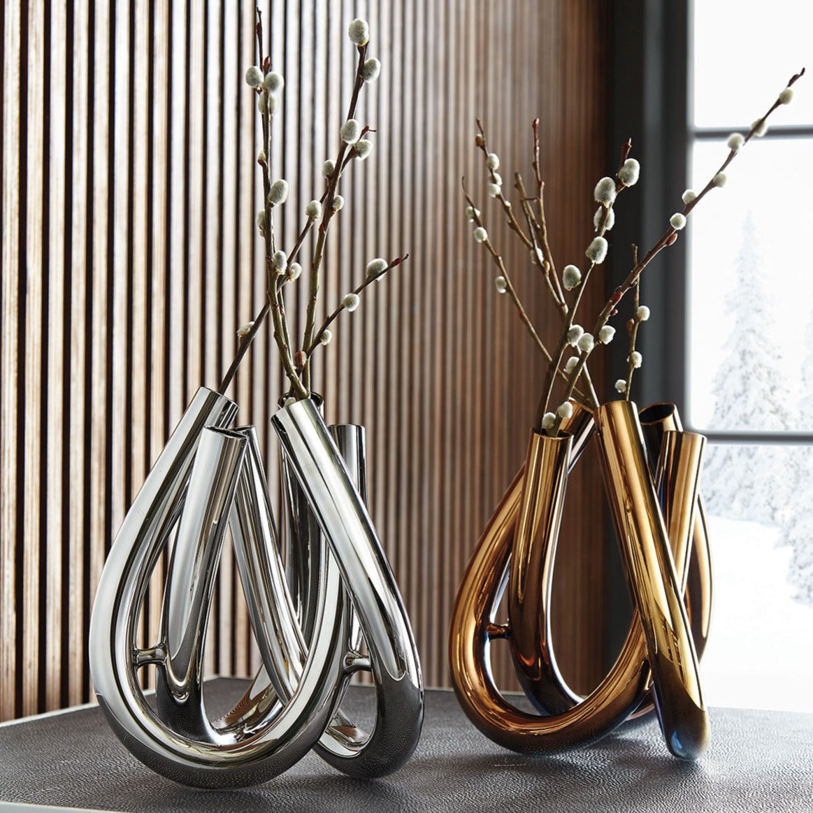 Two modern Trui Vases by Rosenthal—one silver PVD-coated, one gold—hold pussy willow branches. They sit on a dark surface before wood paneling and a window with a snowy outdoor view.
