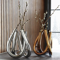 Two modern Trui Vases by Rosenthal—one silver PVD-coated, one gold—hold pussy willow branches. They sit on a dark surface before wood paneling and a window with a snowy outdoor view.