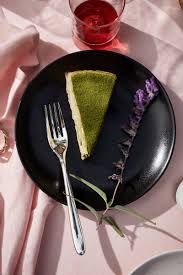 A slice of green matcha cake sits on a L'Objet Terra Iron Dinnerware plate, next to a fork and purple flowers, arranged on a pink tablecloth with a glass of red drink.