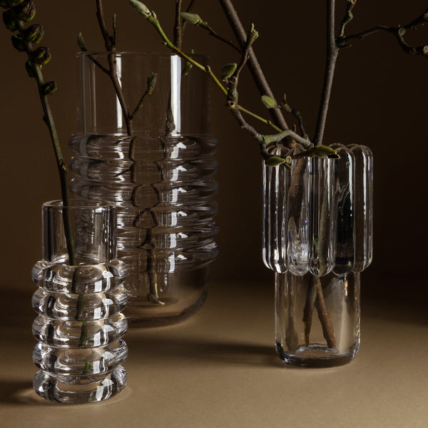 Three Tom Dixon Press Vase Medium pieces, with unique ribbed designs, display bare branches. Arranged on a brown surface against a brown background, they create a minimalist and elegant composition.