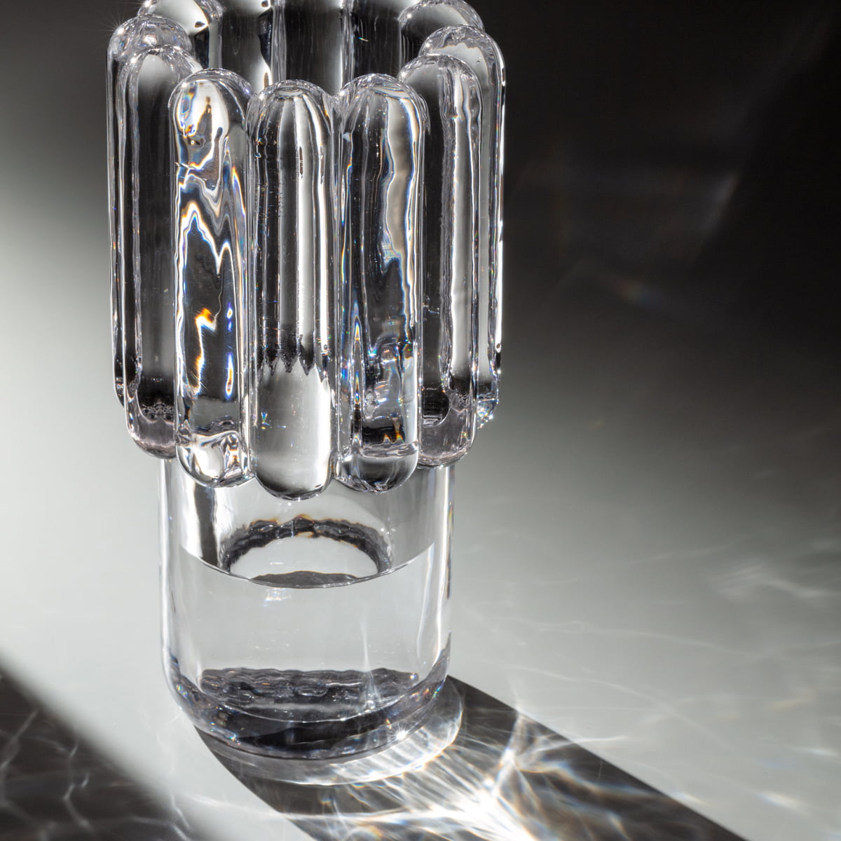 The Press Vase Medium by Tom Dixon, a clear cylindrical glass vase with vertical, rounded ridges, stands on a reflective surface and creates intricate light and shadow patterns.
