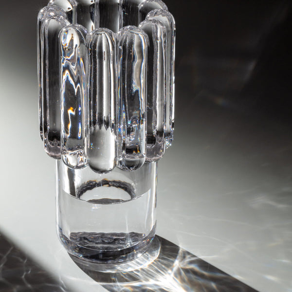 The Press Vase Medium by Tom Dixon, a clear cylindrical glass vase with vertical, rounded ridges, stands on a reflective surface and creates intricate light and shadow patterns.