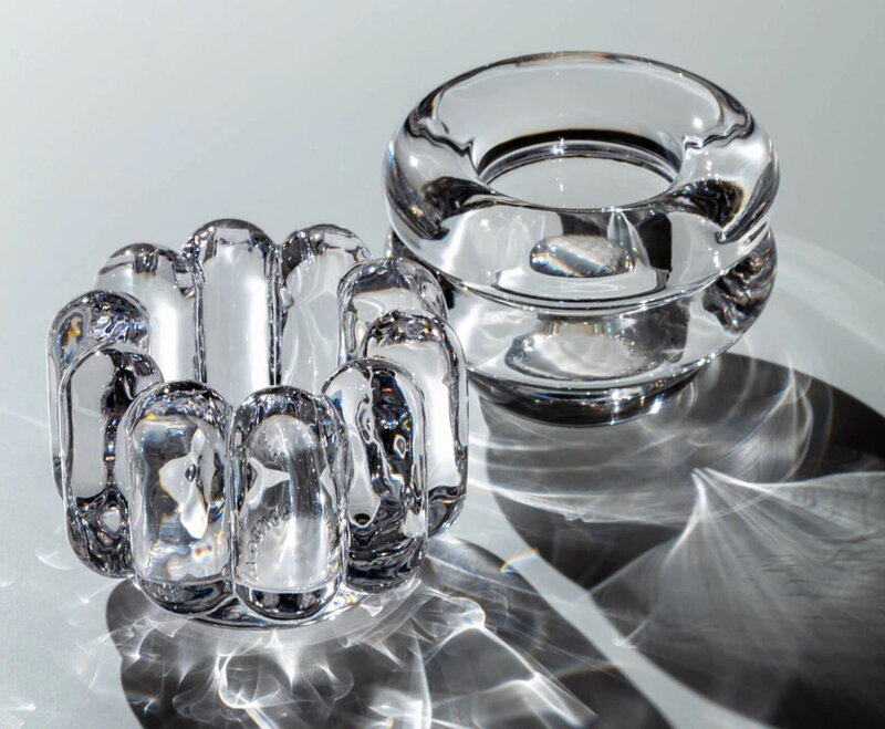 Two clear glass objects rest on a reflective surface, creating intricate shadows. On the left is the Tom Dixon Press Vertical Tealight; on the right, a smooth, circular glass candle holder features a central opening.