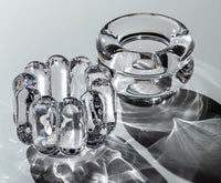 Two clear glass objects rest on a reflective surface, creating intricate shadows. On the left is the Tom Dixon Press Vertical Tealight; on the right, a smooth, circular glass candle holder features a central opening.