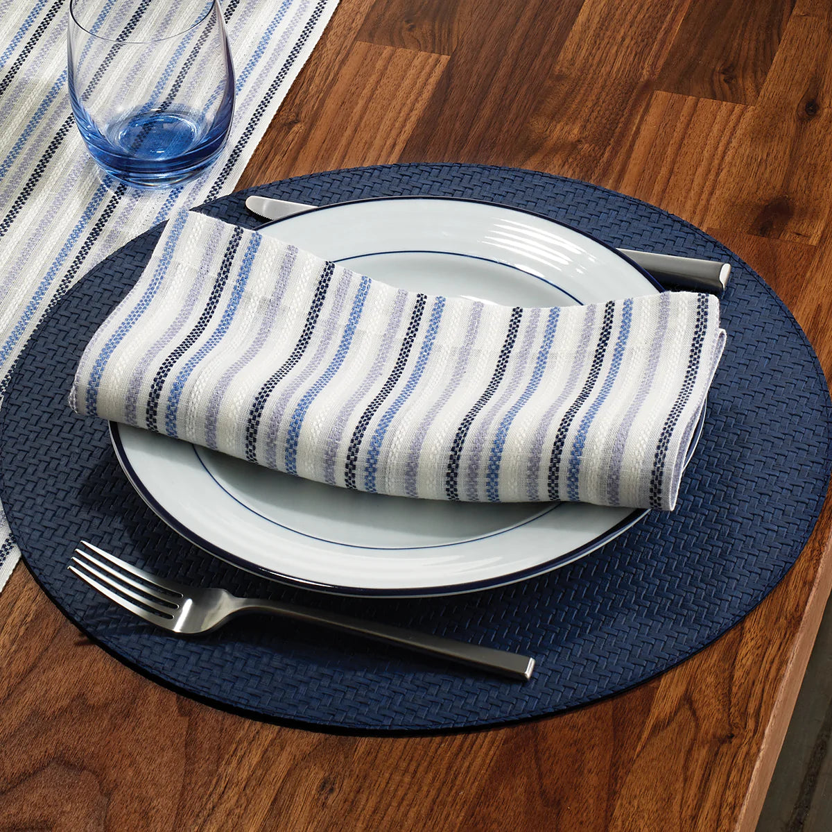 A place setting on a wooden table features Bodrum Wicker Round Placemat Navy, white plates, a striped blue and white napkin, silver fork and knife, and a clear glass with a blue tint. Set includes 4 placemats.