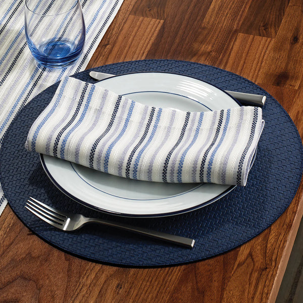 A place setting on a wooden table features Bodrum Wicker Round Placemat Navy, white plates, a striped blue and white napkin, silver fork and knife, and a clear glass with a blue tint. Set includes 4 placemats.