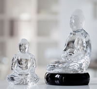 Two Baccarat Little Crystal Buddha statues by Baccarat sit side by side on a reflective surface. The larger figure is displayed on a black base; both feature serene, seated meditation poses and are expertly crafted from clear crystal.