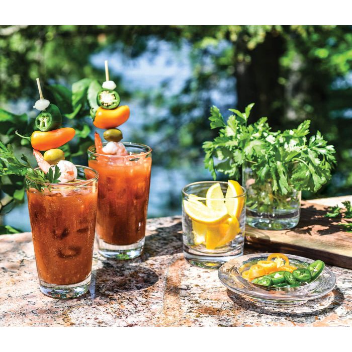 Two Bloody Mary cocktails in Simon Pearce Ascutney Pint glasses are garnished with shrimp, olives, jalapeños, and peppers. Nearby: parsley-filled glasses, lemon wedges, sliced peppers, all set against a lush green backdrop.