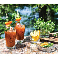 Two Bloody Mary cocktails in Simon Pearce Ascutney Pint glasses are garnished with shrimp, olives, jalapeños, and peppers. Nearby: parsley-filled glasses, lemon wedges, sliced peppers, all set against a lush green backdrop.