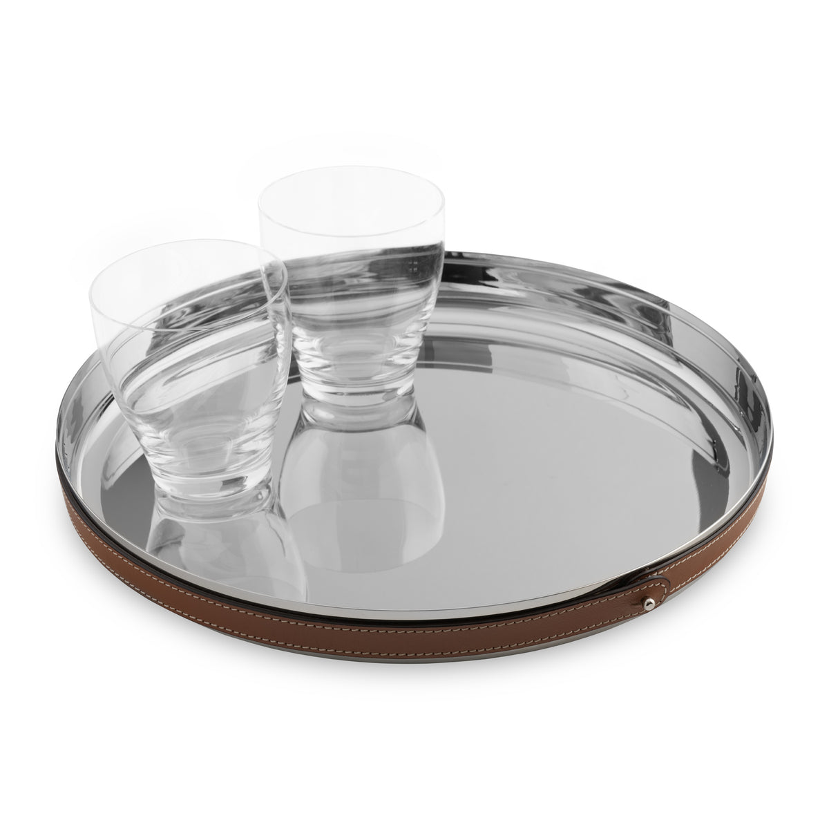 The Nambe Tahoe Bar Tray, a round stainless steel design with a brown leather accent, holds two clear glass tumblers. Both tray and glasses are elegantly mirrored on the shiny surface.