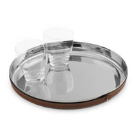The Nambe Tahoe Bar Tray, a round stainless steel design with a brown leather accent, holds two clear glass tumblers. Both tray and glasses are elegantly mirrored on the shiny surface.