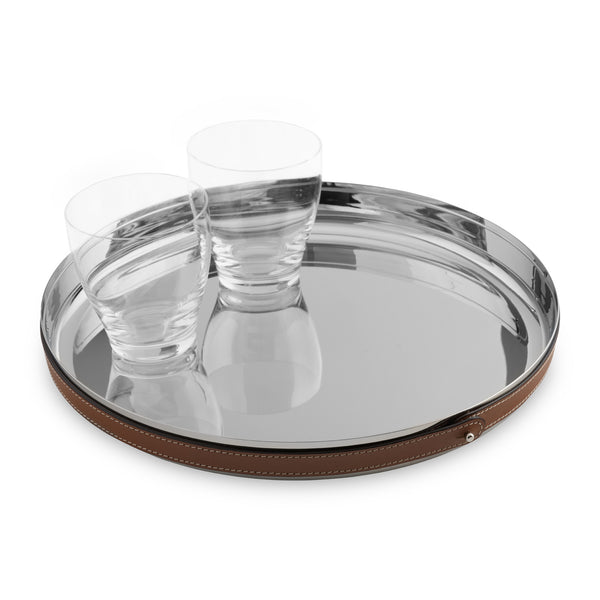 The Nambe Tahoe Bar Tray, a round stainless steel design with a brown leather accent, holds two clear glass tumblers. Both tray and glasses are elegantly mirrored on the shiny surface.