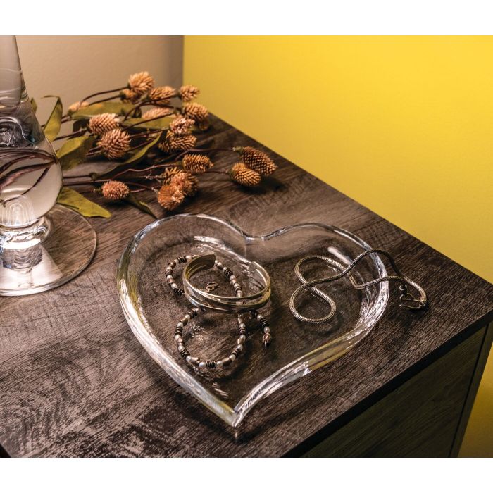 The Simon Pearce Heart Tray, perfect as a sweet gift, displays silver jewelry on a dark wooden surface beside a lamp and dried flowers, illuminated by warm yellow light in the background.