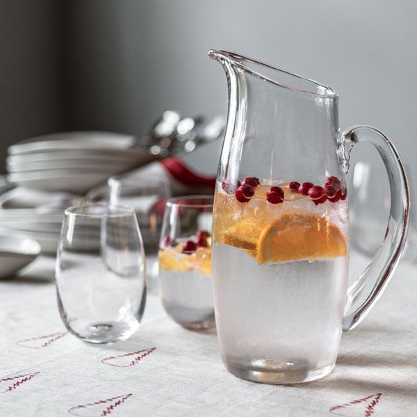 The Simon Pearce Addison Pitcher, filled with fruit-infused water, sits on a table beside empty glasses, stacked plates, and utensils—showcasing the brand's elegant craftsmanship.