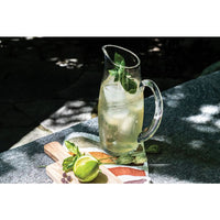 A Simon Pearce Addison Pitcher filled with iced lemonade and mint sits on a stone surface beside a wooden board with sliced lime and fresh mint, partially shaded by dappled sunlight.