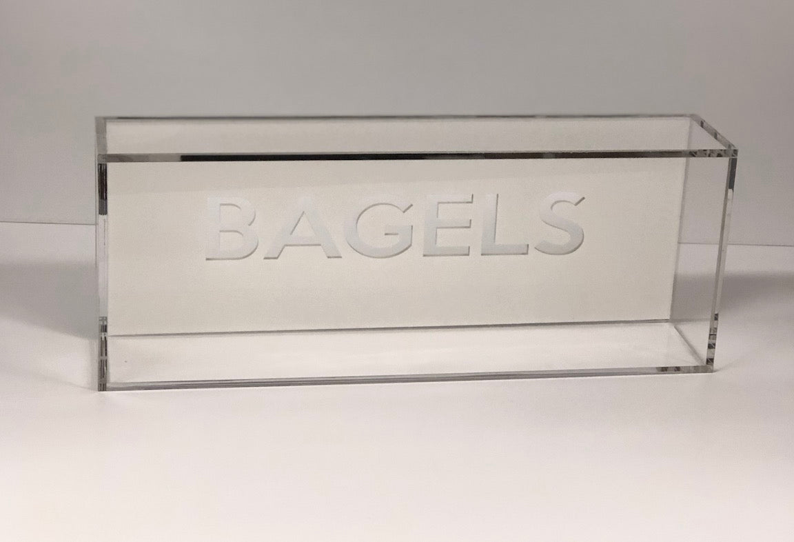 The Eye Candy Bagel Tray is a clear, food-safe acrylic box with "BAGELS" etched in capitals on the front. Its sleek rectangular design is empty and set against a plain white backdrop.