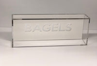 The Eye Candy Bagel Tray is a clear, food-safe acrylic box with "BAGELS" etched in capitals on the front. Its sleek rectangular design is empty and set against a plain white backdrop.