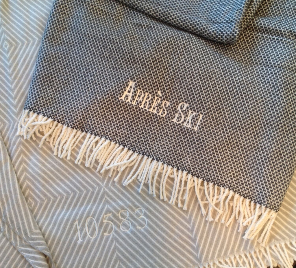 A Soft Idea’s Personalized Throw Blanket in gray with white fringe features “Après Ski” and the number 10583 embroidered in white, displayed on a patterned fabric.