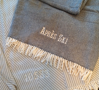 A Soft Idea’s Personalized Throw Blanket in gray with white fringe features “Après Ski” and the number 10583 embroidered in white, displayed on a patterned fabric.