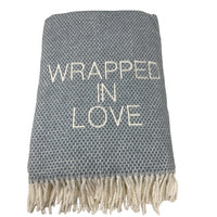 The A Soft Idea Wrapped in Love Throw is a folded gray and white woven blanket with white fringe and "WRAPPED IN LOVE" embroidered on the front. Machine washable and super soft, it’s a perfect holiday gift.