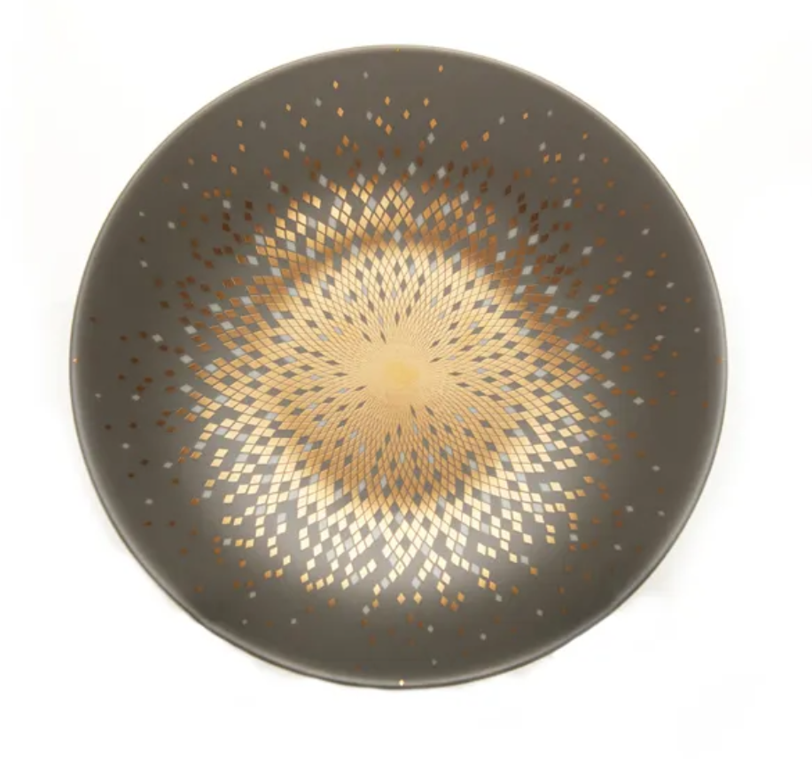 The Souffle D'or Dinnerware by Haviland is a round ceramic plate with a matte dark gray surface and a central geometric gold and beige starburst mosaic. This handmade piece is shown against a white background.