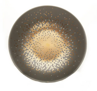 The Souffle D'or Dinnerware by Haviland is a round ceramic plate with a matte dark gray surface and a central geometric gold and beige starburst mosaic. This handmade piece is shown against a white background.