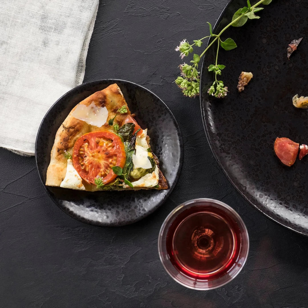 A slice of pizza with tomato, cheese, and basil sits on L'Objet Alchimie Dinnerware Black, next to a glass of red wine, fresh herbs, and an empty plate with crumbs on a dark textured surface.