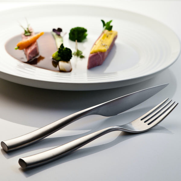 A Mepra Arte Flatware 5 Piece Setting in stainless steel rests on a white surface in the foreground, while a white plate with an artfully arranged gourmet dish appears in the background.