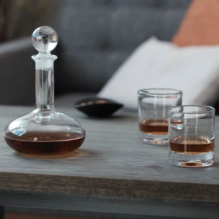 A Simon Pearce Ascutney Double Old Fashioned Set of 2, filled with whiskey, sits on a wooden table next to a glass decanter and stopper. A small black bowl and a sofa with a cushion are visible in the background.