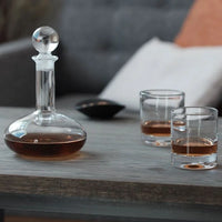 A Simon Pearce Ascutney Double Old Fashioned Set of 2, filled with whiskey, sits on a wooden table next to a glass decanter and stopper. A small black bowl and a sofa with a cushion are visible in the background.