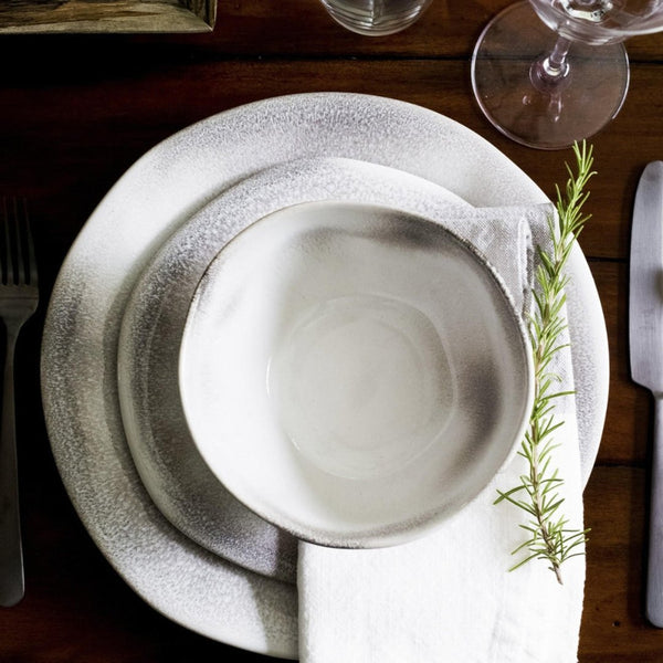 A close-up of a table set with Vietri’s Aurora Dinnerware Ash, featuring a ceramic bowl and plate with subtle gray accents, a cloth napkin with rosemary, elegant cutlery, and a wine glass on a wooden table.
