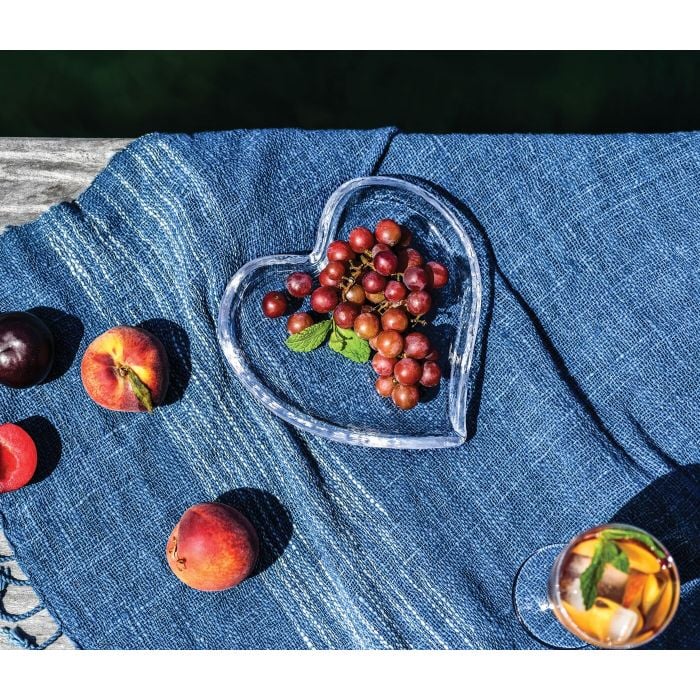 A Simon Pearce Heart Tray holds red grapes atop a blue woven cloth, accompanied by peaches, plums, and a glass of iced tea with mint on rustic wood—a charming gift idea for any occasion.