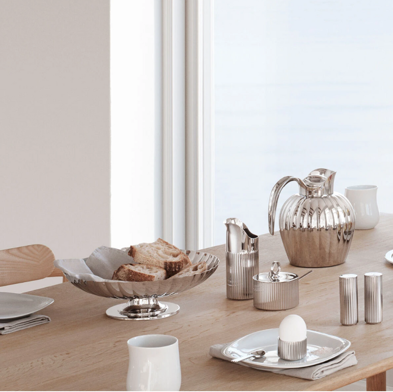 A breakfast table set with silverware featuring a Georg Jensen Bernadotte Stainless Creamer, teapot, sugar bowl, stainless steel milk jug, bread basket of sliced bread, egg cup, plates, cups, and napkins by a large window.