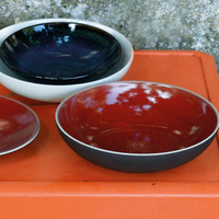 Three round Tourron Dinnerware Cerise ceramic bowls by Jars - Rosenthal sit on an orange surface; two have deep red interiors and one glossy dark blue, each with a pretty glaze. The dishwasher safe bowls are set before a textured stone background.