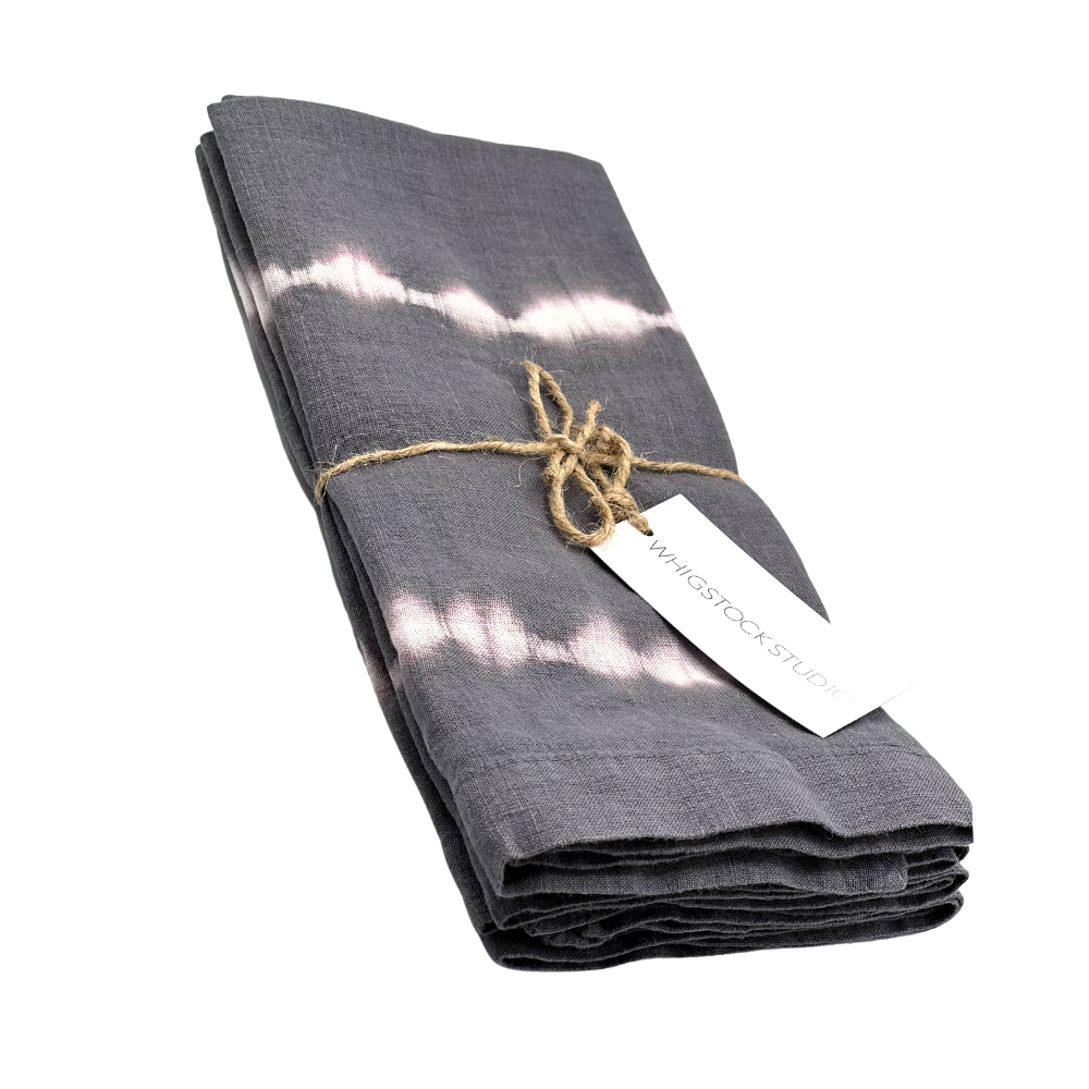 The Hand Dyed Linen Napkin Set of 4 - Midnight by Whigstock Studio features folded, dark gray napkins with white tie-dye stripes, tied with twine and a branded tag, displayed on a white background—ideal for sustainable essentials enthusiasts.