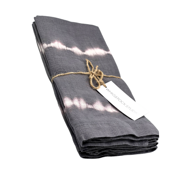 The Hand Dyed Linen Napkin Set of 4 - Midnight by Whigstock Studio features folded, dark gray napkins with white tie-dye stripes, tied with twine and a branded tag, displayed on a white background—ideal for sustainable essentials enthusiasts.