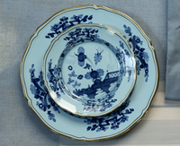 Two Oriente Italiano Dinnerware Iris plates by Ginori 1735, featuring blue and white chinoiserie floral motifs with gold trim and scalloped edges, are stacked on a light blue fabric surface, evoking Italian porcelain elegance.