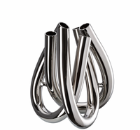 The Trui Vase by Rosenthal is a modern, abstract piece featuring shiny, polished metal tubes with a sleek PVD coating, curving and interlocking at the base as it stands upright on a white background.
