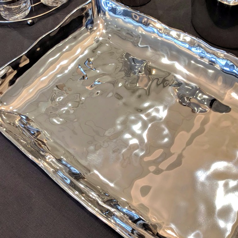 The Beatriz Ball Soho Rectangle Pennsylvania Platter, an elegant aluminum alloy tray with wavy edges and a rippled surface, rests on a dark tablecloth beside glassware and a reflective metal accent for a stylish oven-to-table display.