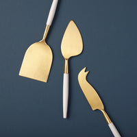 The Be Home White & Gold Cheese Set features three uniquely finished gold-toned knives with white handles—a flat spatula, pointed serving knife, and fork-tipped serrated knife—elegantly arranged on a dark blue background.