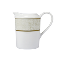 The Sauvage Gold Dinnerware by Bernardaud features a white ceramic pitcher with a handle, a textured beige band like embossed snakeskin, and a thin gold line, all in the elegant style of classic Limoges porcelain.